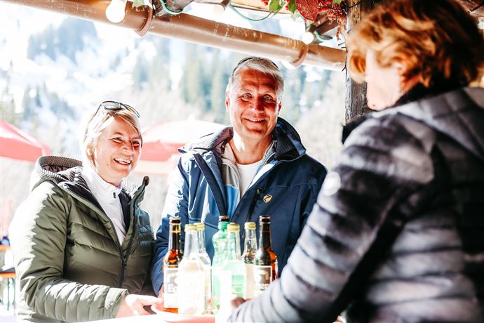 Ein fröhliches Paar steht an einem Bartresen und unterhält sich mit einer Kellnerin. Im Hintergrund sind Berge und Sonnenschirme zu sehen.