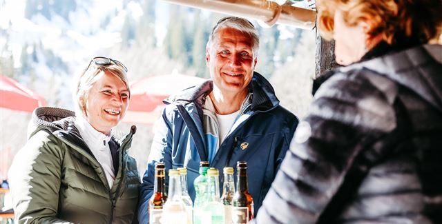 Ein fröhliches Paar steht an einem Bartresen und unterhält sich mit einer Kellnerin. Im Hintergrund sind Berge und Sonnenschirme zu sehen.
