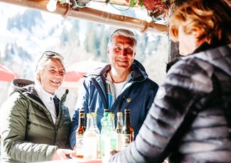 Ein fröhliches Paar steht an einem Bartresen und unterhält sich mit einer Kellnerin. Im Hintergrund sind Berge und Sonnenschirme zu sehen.
