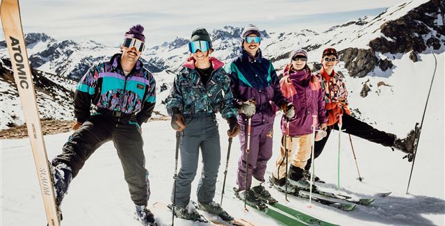 Eine Gruppe von fünf Skifahrern steht auf einer verschneiten Piste in den Bergen. Sie tragen bunte Skibekleidung und halten ihre Skier in den Händen.