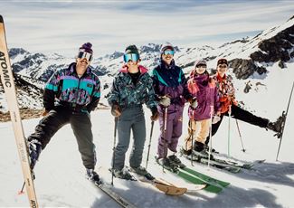 Eine Gruppe von fünf Skifahrern steht auf einer verschneiten Piste in den Bergen. Sie tragen bunte Skibekleidung und halten ihre Skier in den Händen.