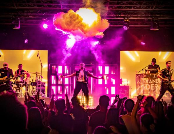 A live band is playing on a stage in the mountains. In the background, there are snow-covered peaks and spectators enjoying the concert.
