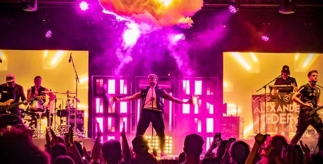 A live band is playing on a stage in the mountains. In the background, there are snow-covered peaks and spectators enjoying the concert.