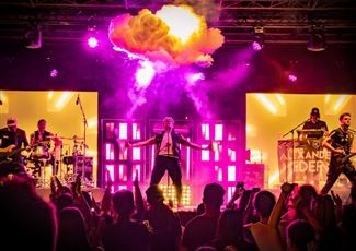 A live band is playing on a stage in the mountains. In the background, there are snow-covered peaks and spectators enjoying the concert.