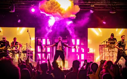 Eine Live-Band spielt auf einer Bühne in den Bergen. Im Hintergrund sind schneebedeckte Gipfel und Zuschauer, die das Konzert genießen.