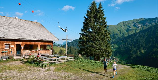 Eine malerische Alm umgeben von grünen Bergen. Zwei Personen joggen auf einem Weg bei klarem Himmel.