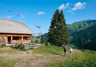Eine malerische Alm umgeben von grünen Bergen. Zwei Personen joggen auf einem Weg bei klarem Himmel.