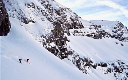 Zwei Skifahrer fahren auf einer schneebedeckten Piste in den Bergen. Die Landschaft ist beeindruckend und zeigt hohe Felsen und frischen Schnee.