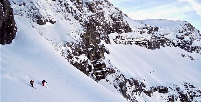 Two skiers are skiing on a snow-covered slope in the mountains. The landscape is impressive and features high cliffs and fresh snow.
