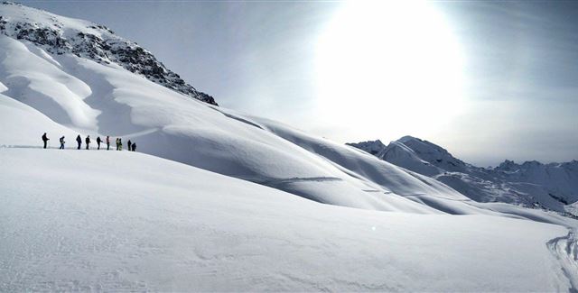 A group of people is hiking over snow-covered hills in an impressive winter landscape. The sun is shining brightly in the sky and illuminating the surroundings.