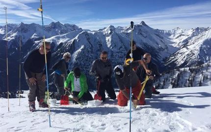 Eine Gruppe von Menschen arbeitet im Schnee auf einem Berg. Im Hintergrund sind majestätische Berggipfel und ein klarer Himmel sichtbar.