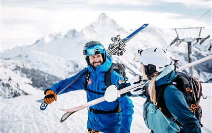 Zwei Skifahrer stehen fröhlich im Schnee und halten ihre Skier in der Hand. Im Hintergrund sind schneebedeckte Berge und ein Skilift zu sehen.