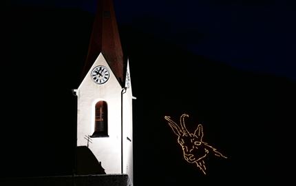 Eine weiß gestrichene Kirche mit einem roten Dach und einer Uhr. Neben der Kirche ist ein beleuchtetes Geweihsymbol zu sehen.