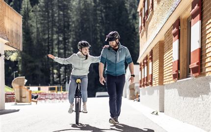 Ein Mann geht neben einem Mädchen, das auf einem Einrad fährt. Beide tragen Helme und lachen in einer sonnigen, natürlichen Umgebung.