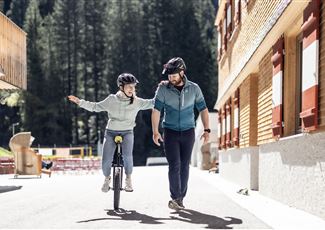 Ein Mann geht neben einem Mädchen, das auf einem Einrad fährt. Beide tragen Helme und lachen in einer sonnigen, natürlichen Umgebung.