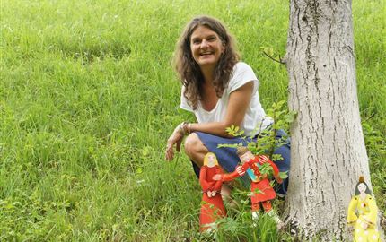 Eine lächelnde Frau sitzt neben einem Baum und posiert. Im Vordergrund sind bunte Figuren auf dem grünen Gras zu sehen.