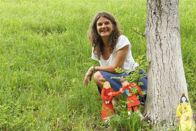 Eine Frau sitzt lächelnd neben einem Baum auf einer grünen Wiese. Um sie herum sind bunte Figuren aufgestellt.