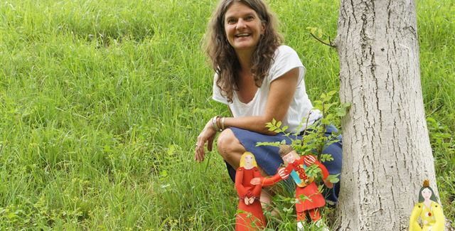 Eine Frau sitzt lächelnd neben einem Baum auf einer grünen Wiese. Um sie herum sind bunte Figuren aufgestellt.