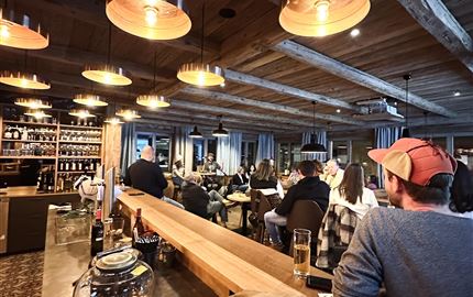 Eine gemütliche Bar mit Holzdekor und warmen Lichtern. Menschen genießen Getränke und unterhalten sich in einer einladenden Atmosphäre.
