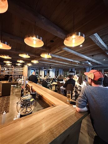 Eine gemütliche Bar mit Holzdekor und warmen Lichtern. Menschen genießen Getränke und unterhalten sich in einer einladenden Atmosphäre.