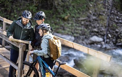 Drei Radfahrer stehen auf einer Brücke und schauen auf eine Karte. Es ist eine grüne, natürliche Umgebung mit einem plätschernden Wasserlauf.