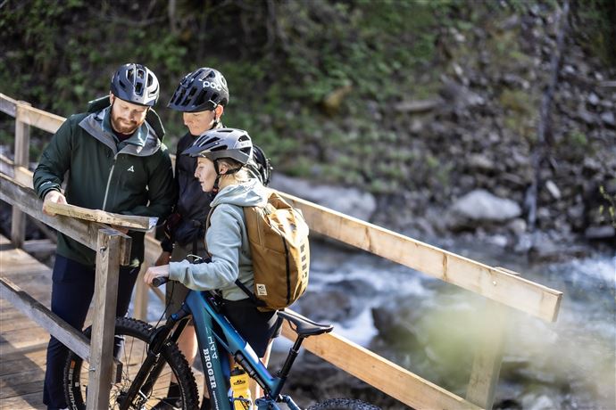 Drei Radfahrer stehen auf einer Brücke und schauen auf eine Karte. Es ist eine grüne, natürliche Umgebung mit einem plätschernden Wasserlauf.