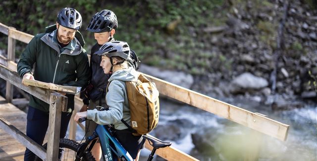 Drei Radfahrer stehen auf einer Brücke und schauen auf eine Karte. Es ist eine grüne, natürliche Umgebung mit einem plätschernden Wasserlauf.