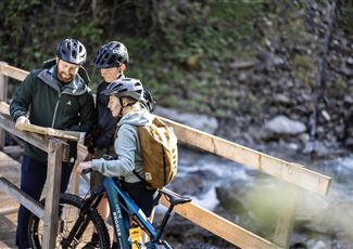 Drei Radfahrer stehen auf einer Brücke und schauen auf eine Karte. Es ist eine grüne, natürliche Umgebung mit einem plätschernden Wasserlauf.