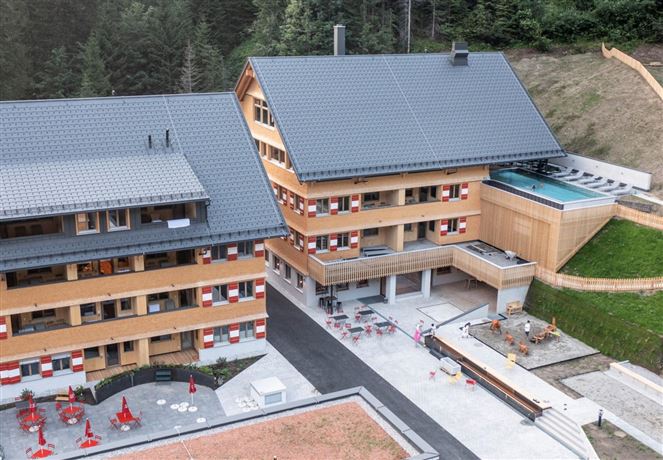 Ein modernes Gebäude im alpinen Stil, umgeben von Bäumen. Im Vordergrund befindet sich ein Bereich mit Sitzgelegenheiten und einem Außenpool.