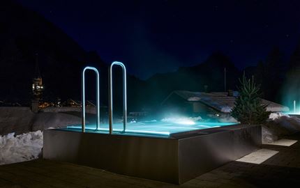 Ein modernes, beleuchtetes Schwimmbad in den Bergen bei Nacht. Umgeben von Schnee und dekorativen Lichtern bietet es eine entspannende Atmosphäre.