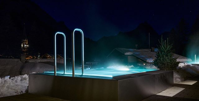 Ein modernes, beleuchtetes Schwimmbad in den Bergen bei Nacht. Umgeben von Schnee und dekorativen Lichtern bietet es eine entspannende Atmosphäre.