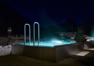 Ein modernes, beleuchtetes Schwimmbad in den Bergen bei Nacht. Umgeben von Schnee und dekorativen Lichtern bietet es eine entspannende Atmosphäre.
