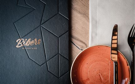 Ein elegantes Restaurantmenü in Schwarz mit goldener Schrift. Daneben ein orangefarbener Teller und Besteck auf einem weißen Serviette.