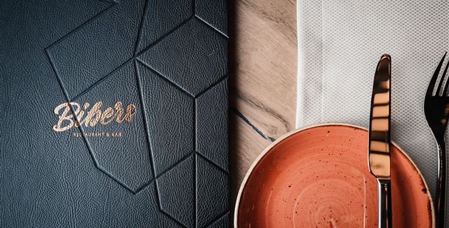 Ein elegantes Restaurantmenü in Schwarz mit goldener Schrift. Daneben ein orangefarbener Teller und Besteck auf einem weißen Serviette.