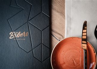 Ein elegantes Restaurantmenü in Schwarz mit goldener Schrift. Daneben ein orangefarbener Teller und Besteck auf einem weißen Serviette.