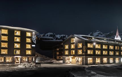 Ein modernes Hotelgebäude bei Nacht, umgeben von schneebedeckten Bergen. Die Fenster leuchten warm und einladend im Dunkeln.