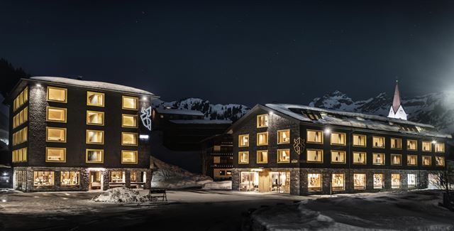 Ein modernes Hotelgebäude bei Nacht, umgeben von schneebedeckten Bergen. Die Fenster leuchten warm und einladend im Dunkeln.
