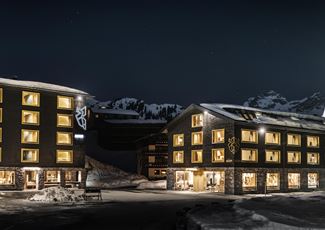 Ein modernes Hotelgebäude bei Nacht, umgeben von schneebedeckten Bergen. Die Fenster leuchten warm und einladend im Dunkeln.