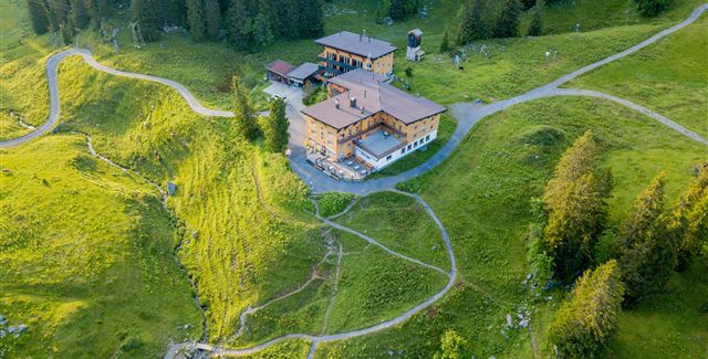 Eine malerische Landschaft mit einem großen Gebäude umgeben von grünen Wiesen und Wäldern. Wanderwege führen durch die sanfte Hügellandschaft.