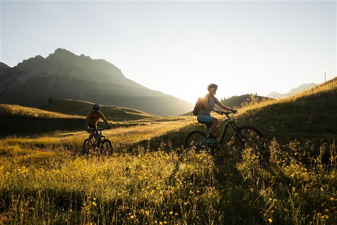Zwei Radfahrer fahren durch eine blühende Wiese in den Bergen. Die Sonne scheint und die Landschaft ist wunderschön.