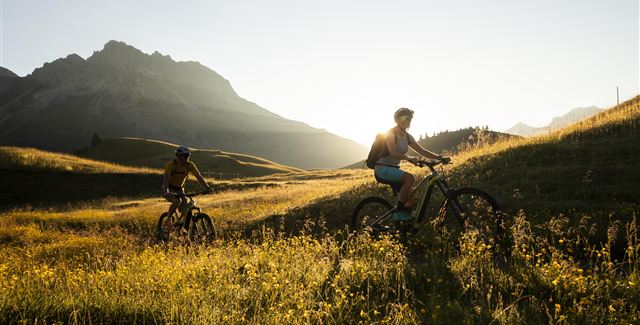 Zwei Radfahrer fahren durch eine blühende Wiese in den Bergen. Die Sonne scheint und die Landschaft ist wunderschön.