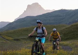 Zwei Radfahrer erkunden eine malerische Landschaft mit grünen Wiesen und blühenden Blumen. Im Hintergrund sind Berge unter einem klaren Himmel zu sehen.