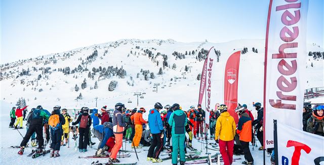 A group of skiers gathers on a snowy slope. In the background, mountains and flags can be seen.
