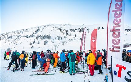Eine Gruppe von Skifahrern versammelt sich auf einem verschneiten Hang. Im Hintergrund sind Berge und Fahnen zu sehen.