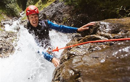 Ein Abenteurer in einem roten Helm klettert an einem nassen Felsen entlang, während Wasser spritzt. Die Umgebung ist felsig und grün.