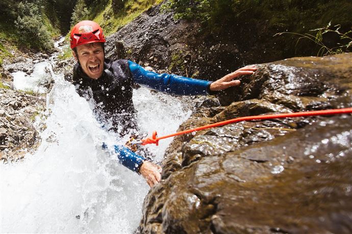 Ein Abenteurer in einem roten Helm klettert an einem nassen Felsen entlang, während Wasser spritzt. Die Umgebung ist felsig und grün.