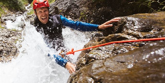 Ein Abenteurer in einem roten Helm klettert an einem nassen Felsen entlang, während Wasser spritzt. Die Umgebung ist felsig und grün.
