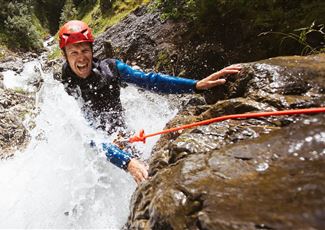 Ein Abenteurer in einem roten Helm klettert an einem nassen Felsen entlang, während Wasser spritzt. Die Umgebung ist felsig und grün.