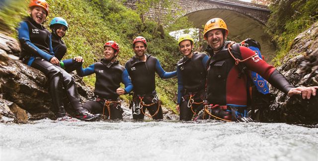 Sechs Personen in Neoprenanzügen stehen im Wasser eines Flusses und lächeln in die Kamera. Im Hintergrund ist eine Brücke und üppiges Grün zu sehen.