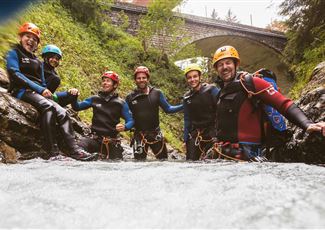 Sechs Personen in Neoprenanzügen stehen im Wasser eines Flusses und lächeln in die Kamera. Im Hintergrund ist eine Brücke und üppiges Grün zu sehen.